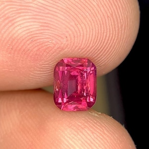 May include: A close-up of a radiant, rectangular-cut, pink gemstone. The gem is held between two fingers, showcasing its vibrant color and facets. The gemstone's shape is a rectangle with cut corners, reflecting light beautifully.