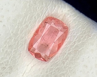 Pode incluir: Uma pedra preciosa facetada, de forma retangular, com um tom rosa suave. A gema tem um corte almofada com múltiplas facetas que refletem a luz. A pedra está sobre um fundo branco texturizado.