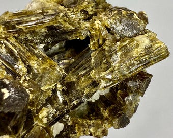 Może przedstawiać: Zbliżenie na okaz minerału z warstwowymi, półprzezroczystymi, oliwkowozielonymi kryształami. Kryształy mają prążkowany wygląd i są poprzetykane białymi i ciemnobrązowymi sekcjami. Ogólna struktura jest złożona i poszarpana.