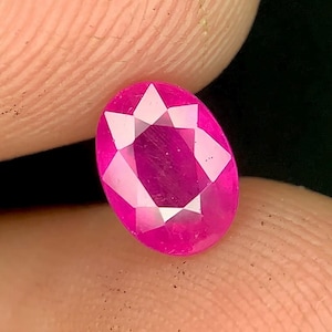 Può includere: Una gemma rosa brillante, sfaccettata, di forma ovale. La gemma ha un motivo a forma di stella al centro ed è tenuta tra le dita. Le sfaccettature della gemma riflettono la luce, creando un effetto scintillante. Lo sfondo è scuro.
