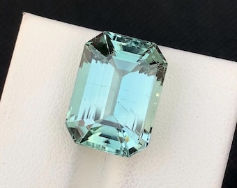 May include: A rectangular-cut, aquamarine gemstone with a light blue-green hue. The gem is faceted, showcasing internal reflections and a polished surface. It rests on a white surface against a black backdrop.