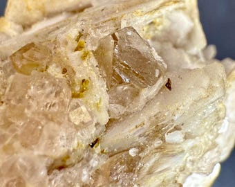 Może przedstawiać: Zbliżenie na okaz minerału z krystalicznymi strukturami w odcieniach beżu i brązu. Kryształy są przezroczyste, z widocznymi wewnętrznymi prążkami. Otaczająca matryca ma jaśniejszy, białawy kolor i lekko szorstką teksturę.