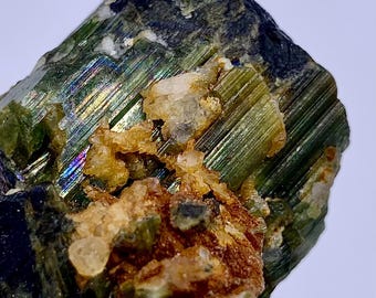 May include: Close-up of a raw mineral specimen. The stone has a dark green, striated surface with iridescent flashes. Patches of tan and clear crystalline formations are visible, creating a textured appearance. A natural, uncut gemstone.