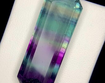 May include: A rectangular, faceted fluorite gemstone with a gradient of colours. The stone transitions from deep purple at the base to shades of green and blue at the top. The gemstone is displayed on a white surface.