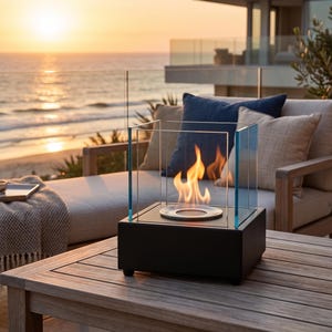 May include: A contemporary black tabletop fireplace with a glass surround. Flames are visible within the glass, set on a wooden table. The background shows a beach scene with the sea and sunset. The fireplace is approximately 30 cm square.