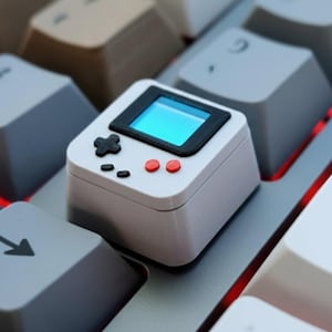 May include: A white, miniature retro game console keycap with a black screen and red buttons sits on a computer keyboard. The keycap has a D-pad and two small buttons. The keyboard keys are various shades of gray and white.