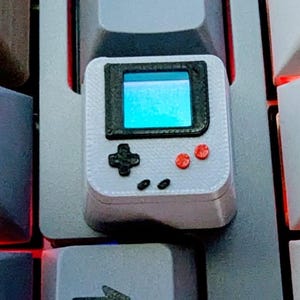May include: A custom keycap shaped like a miniature handheld gaming device. The keycap is light gray with a black screen and red buttons. The directional pad and other buttons are black. The keycap is designed to fit on a mechanical keyboard.