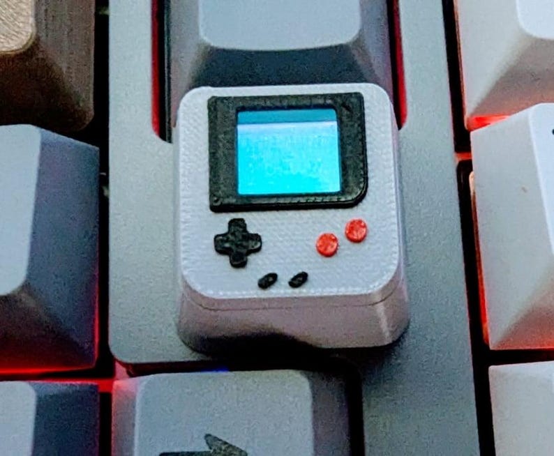 May include: A custom keycap shaped like a miniature handheld gaming device. The keycap is light gray with a black screen and red buttons. The directional pad and other buttons are black. The keycap is designed to fit on a mechanical keyboard.