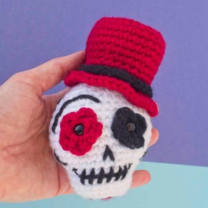 Può includere: Un teschio all'uncinetto con un cappello a cilindro rosso, accenti neri e dettagli floreali per gli occhi. Il teschio è bianco con cuciture nere per la bocca e i contorni degli occhi. Il cappello ha una fascia nera.