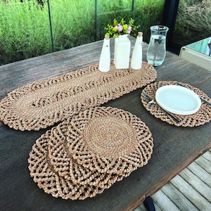 May include: A woven table runner and placemats set. The runner is oval-shaped with a decorative border. The placemats are round with a similar border. The set is made of a natural fiber in a light brown color. A white plate and silverware are on one of the placemats.