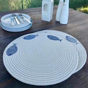 Op de afbeelding: Twee ronde, crèmekleurige placemats met een spiraalontwerp en blauwe visillustraties. De placemats liggen op een houten tafel, met een stapel borden, bestek en decoratieve witte keramische stukken op de achtergrond.