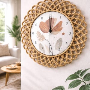 Peut inclure: Une horloge murale ronde avec un cadre tissé de couleur naturelle. Le cadran de l'horloge présente un motif floral dans des tons pêche et beige, avec des aiguilles et des chiffres noirs. L'horloge est fixée sur un mur blanc.