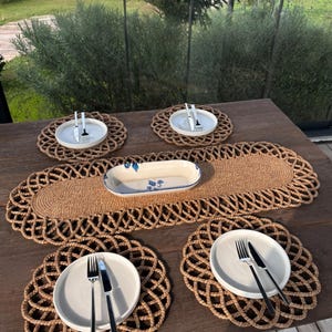 Könnte beinhalten: Ein gedeckter Tisch mit gewebten Jute-Tischsets und einem Tischläufer. Jede Gedeck enthält einen weißen Teller, eine Gabel und ein Messer. Eine rechteckige Servierschale mit floralem Design befindet sich in der Mitte.