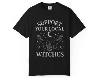 May include: Black t-shirt with the text "SUPPORT YOUR LOCAL WITCHES" in a stylized font. The design includes hands reaching up towards a crescent moon, stars, and a crystal. A casual tee for those interested in witchcraft.