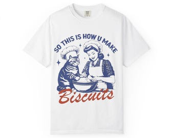 Puede incluir: Camiseta blanca con un diseño gráfico de estilo retro. El diseño presenta a una mujer y un gato, ambos con gorros de chef, mezclando ingredientes en un cuenco. El texto dice "SO THIS IS HOW U MAKE Biscuits" en azul y rojo.