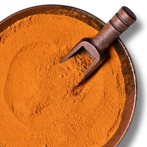 Peut inclure: Gros plan sur un bol en céramique marron rempli d'une poudre orange vif. Une petite cuillère en bois sombre repose à l'intérieur du bol, partiellement immergée dans la poudre. Le bol est posé sur fond blanc.