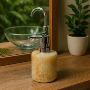 May include: A decorative soap dispenser with a silver pump, made of yellow and gold-veined stone. The dispenser sits on a wooden surface, with a glass sink and mirror in the background. A small potted plant is also visible.