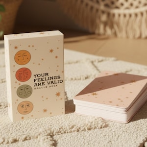 Op de afbeelding: Een orakelkaartspel met een crèmekleurige doos. De doos bevat illustraties van gezichten en de tekst "YOUR FEELINGS ARE VALID ORACLE DECK." De kaarten zijn roze met gouden sterren. Het spel is ontworpen voor spirituele begeleiding.