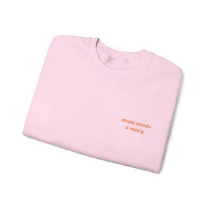 May include: A light pink sweatshirt with the text "mum needs a marg" in orange lettering. The crew neck sweatshirt is folded neatly, showcasing the text on the front. The casual top is made of a soft material.