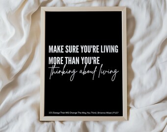 May include: A framed black print with white text that reads "MAKE SURE YOU'RE LIVING MORE THAN YOU'RE thinking about living." The print is in a light wood frame. Below the quote is the text "101 Essays That Will Change The Way You Think | Brianna Wiest | P107."