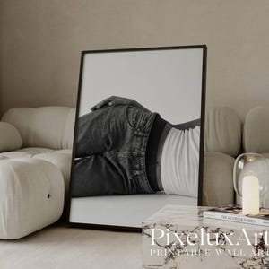 May include: Monochrome framed print depicting a person in denim jeans and a white top. The artwork is in a contemporary living room with neutral colours, a buttoned ottoman, and a circular rug. The print is leaning against a wall.