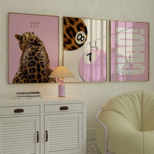 Op de afbeelding: Drie ingelijste kunstprints met roze achtergronden. Eén toont een luipaard, een andere een achtbal en de derde zegt "OHIO!". Een kleine lamp en een witte kast staan voor de kunst. De kunst is in goud omlijst.