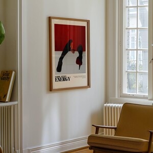 May include: A framed poster with a red and white background and black figures. The poster has the word "ENERGY" in bold letters. The poster is hanging on a white wall. A mustard-colored armchair sits below the poster. A large green plant is visible.
