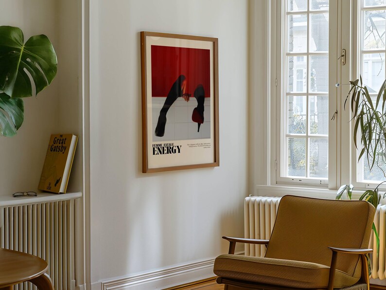 May include: A framed poster with a red and white background and black figures. The poster has the word "ENERGY" in bold letters. The poster is hanging on a white wall. A mustard-colored armchair sits below the poster. A large green plant is visible.