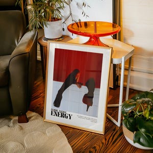 May include: A framed art print with a gold border, featuring a red and white graphic with the words "FEMME FATALE ENERGY". The artwork depicts a figure in a red high heel shoe and black clothing. The print is displayed on a wooden floor.
