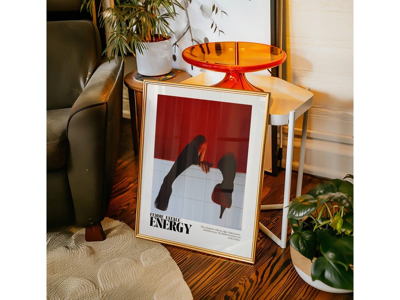 May include: A framed art print with a gold border, featuring a red and white graphic with the words "FEMME FATALE ENERGY". The artwork depicts a figure in a red high heel shoe and black clothing. The print is displayed on a wooden floor.