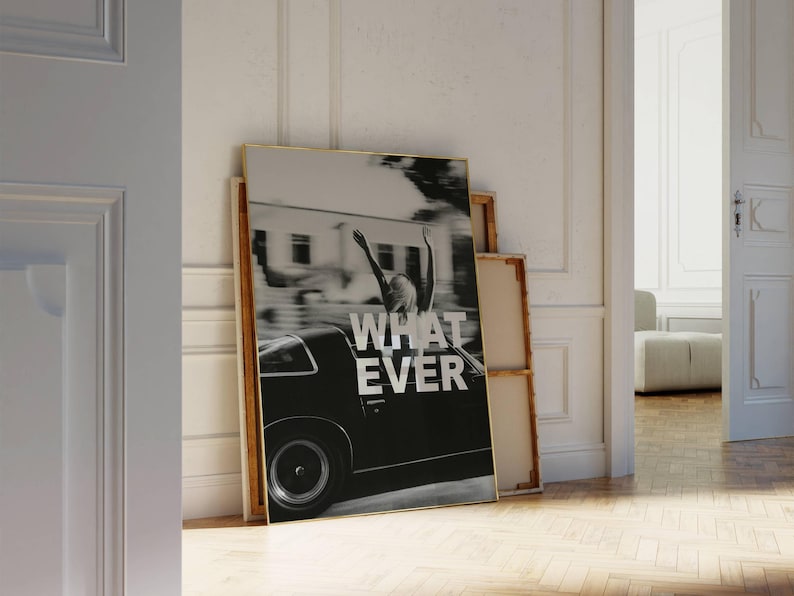 May include: Monochrome framed print showing a person with arms raised from a classic car, with the word "WHATEVER" in white. The artwork is propped against a wall, alongside other framed canvases.