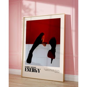 May include: Framed art print featuring a red and white abstract design with black silhouettes of legs and high heels. The text "FEMME FATALE ENERGY" is displayed in bold, black font. The artwork is displayed in a room with pink walls.