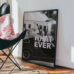 May include: Monochrome framed print showing a classic car and a person with raised arms, with the words "WHAT EVER". A black butterfly chair and a patterned blanket are in the foreground. The print is leaning against a white wall.