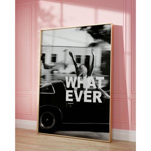 May include: Monochrome framed print showing a person with raised arms in a vintage car. The words "WHATEVER" are in white across the image. The print has a gold frame and is displayed against a pink wall, with a wooden floor.