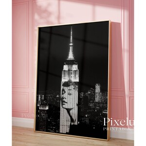 May include: Black and white framed print featuring the Empire State Building with a woman's face superimposed over the building. The print has a gold frame and is displayed against a pink wall. The text "Pixelu Printable" is visible in the bottom right corner.