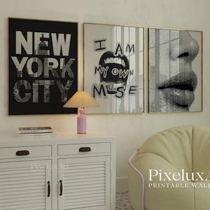 May include: Three framed wall art prints. The first is black and white with "NEW YORK CITY" text. The second says "I AM MY OWN MUSE". The third is a close-up of a face. A pink lamp sits on a white cabinet.