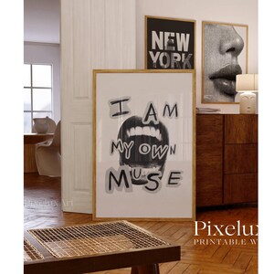May include: A framed print with the text "I AM MY OWN MUSE" above a grayscale image of open lips. The artwork is displayed in a room with other framed art and a wooden cabinet. The room has hardwood floors.