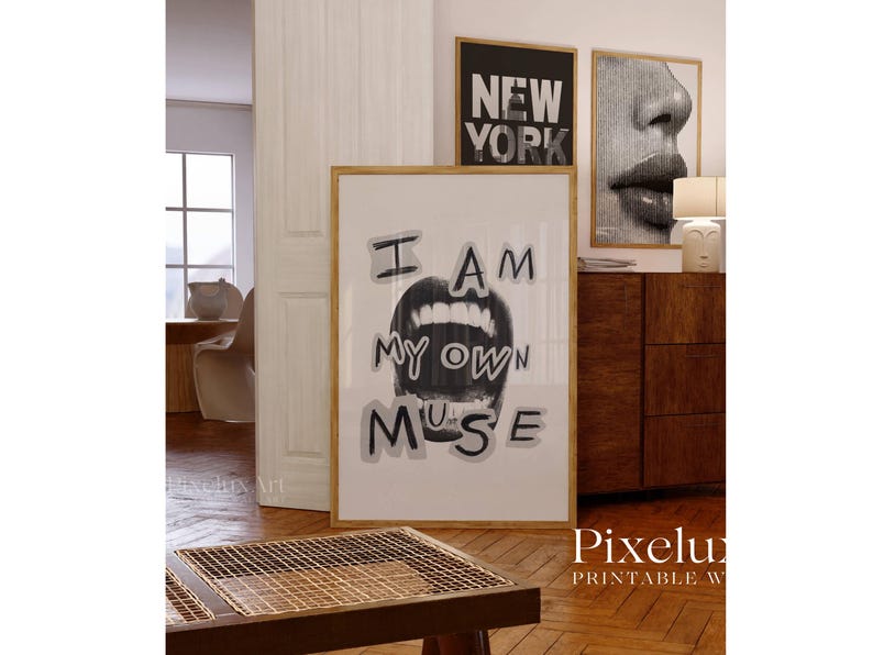 May include: A framed print with the text "I AM MY OWN MUSE" above a grayscale image of open lips. The artwork is displayed in a room with other framed art and a wooden cabinet. The room has hardwood floors.