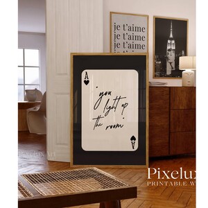 May include: A framed print featuring a playing card design with the text "you light up the room" in elegant script. The ace of hearts and spades are visible. Additional framed prints with French text and a black and white photo are in the background.