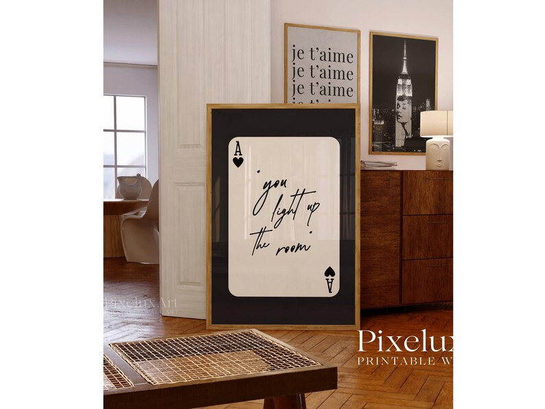 May include: A framed print featuring a playing card design with the text "you light up the room" in elegant script. The ace of hearts and spades are visible. Additional framed prints with French text and a black and white photo are in the background.
