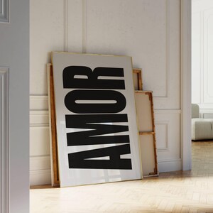 May include: A large framed print with the word "AMOR" in bold black letters against a white background. The print leans against a white wall, with a wooden floor in the foreground. The word "AMOR" means "love" in Spanish and Portuguese.