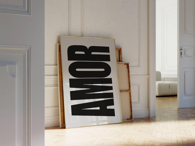 May include: A large framed print with the word "AMOR" in bold black letters against a white background. The print leans against a white wall, with a wooden floor in the foreground. The word "AMOR" means "love" in Spanish and Portuguese.
