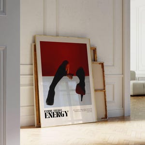 May include: A framed art print with the words "FEMME FATALE ENERGY" in black text. The artwork features a red background with a white horizontal line and a silhouette of legs and high heels. The print is leaning against a white wall.
