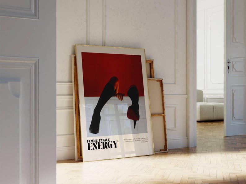 May include: A framed art print with the words "FEMME FATALE ENERGY" in black text. The artwork features a red background with a white horizontal line and a silhouette of legs and high heels. The print is leaning against a white wall.
