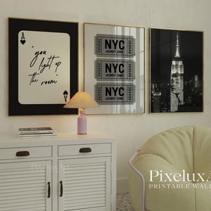 May include: Three framed wall art prints. One features a playing card with the text "you light up the room". Another shows NYC tickets, and the third is a black and white image of the Empire State Building. A small lamp sits on a white cabinet.