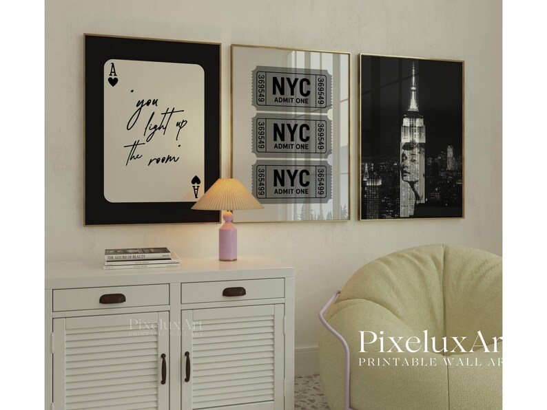 May include: Three framed wall art prints. One features a playing card with the text "you light up the room". Another shows NYC tickets, and the third is a black and white image of the Empire State Building. A small lamp sits on a white cabinet.