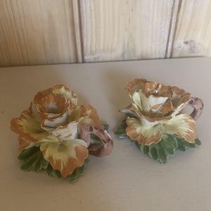May include: Two ceramic teacups shaped like roses. The cups feature cream, orange, and brown petals, with green leaves at the base. The handles are a muted pink. The cups are displayed on a light surface. A decorative floral theme.