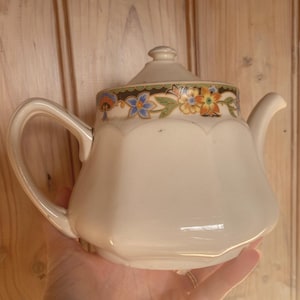 May include: A cream-coloured ceramic teapot with a scalloped body and a matching lid. The upper rim features a decorative band with a repeating pattern of blue, orange, and yellow flowers, green leaves, and black accents on a white background.