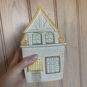 May include: A ceramic house-shaped canister with a removable roof lid. The structure features a white brick-textured facade, a yellow awning over the door, two arched green-trimmed windows on the upper level, and a square brown-trimmed window.