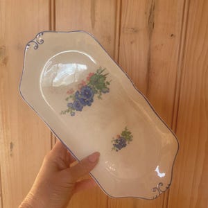 May include: An elongated, cream-coloured ceramic serving tray with a scalloped edge and a thin blue rim. The tray features two floral motifs with blue and pink flowers and green leaves, and decorative blue scrollwork accents at both ends.